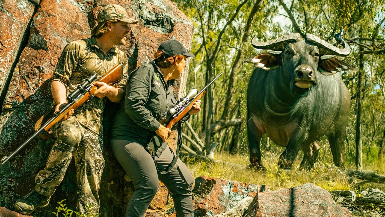 Chris Waters and Lynda Johnson with rifles in their hands crouched behind a rock looking at an angry Water Buffalo